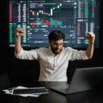 Businessman celebrates stock market success with hands raised in excitement at a trading desk.