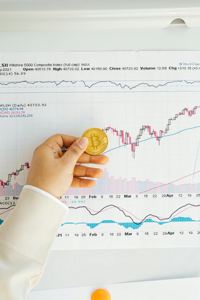 Close-up of a hand holding a Bitcoin coin in front of a stock market chart, symbolizing cryptocurrency trading.